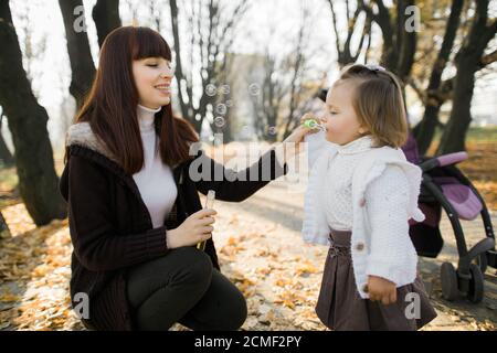 Close up portrait of happy mom and child baby girl, having fun in beautiful autumn park at sunny day, playing, smiling, litlle girl is blowing soap Stock Photo