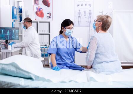 Cardiologist assistant listening heart of senior woman with stethoscope during consultation and wearing face mask against coronavirus outbreak. Medical examination for infections, disease. Stock Photo
