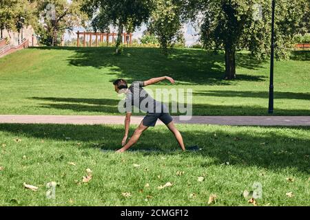 Young athletic man in sportswear doing yoga in the park. Practice triangle asana pose outdoors. People exercising on green grass with yoga mat Stock Photo