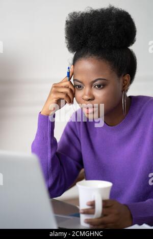Focused Afro-American millennial student woman with afro hairstyle listening lecture, watching webinar on laptop, study online, preparing for exam, ho Stock Photo