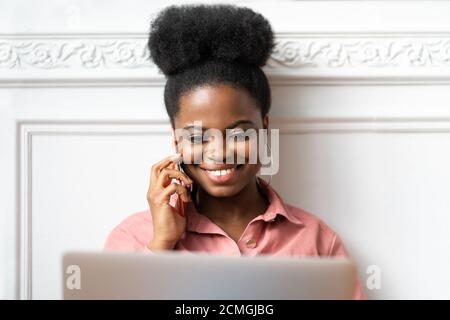 Close up portrait of African American millennial woman with afro hairstyle in pink shirt smiling, talking on phone, looking at laptop. Female employee Stock Photo