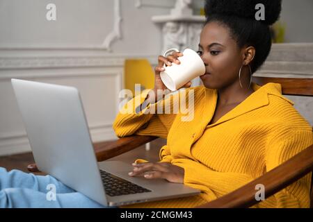 African American biracial woman with afro hairstyle in yellow cardigan sitting on armchair, remote studying, working online on laptop, watching videos Stock Photo