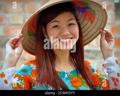 Beautiful young Vietnamese woman with long hair holds the brim of her Asian conical hat with both hands and smiles at camera. Stock Photo