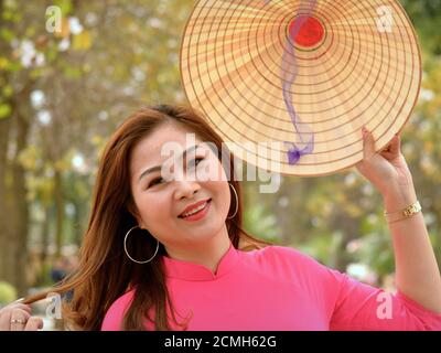 Beautiful young smiling Vietnamese woman holds a traditional Asian conical straw hat with her left hand and poses for the camera. Stock Photo