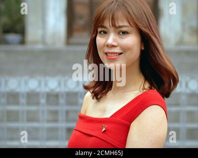 Beautiful young smiling Vietnamese business woman with shoulder-long brown hair wears a self tailored red dress and looks at the camera. Stock Photo