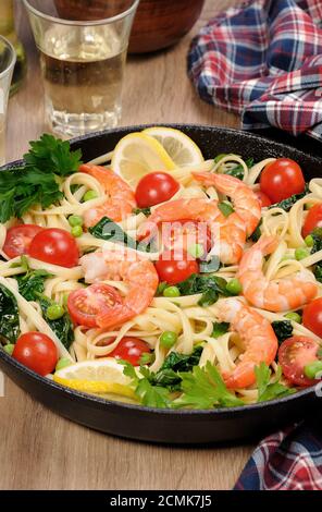 Pasta with fried prawns, peas, tomatoes and spinach in a frying pan, on a table with cider glasses. Stock Photo