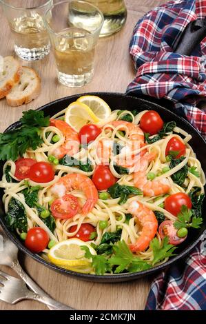 Pasta with fried prawns, peas, tomatoes and spinach in a frying pan, on a table with cider glasses. Stock Photo