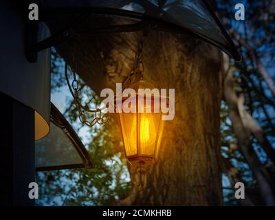 Retro style lantern shines with warm yellow light at dusk. Vintage lantern hangs on a chain under the roof outside the building in a park or forest. Stock Photo