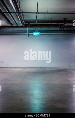 The green illuminated emergency exit notice hangs under the ceiling in an empty underground car park in front of a concrete wall. Stock Photo