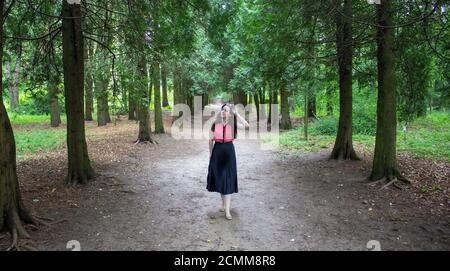 Young girl in sunglasses stands on the path between the rows of fir trees. A girl dressed in a pink blouse and a black skirt. Stock Photo