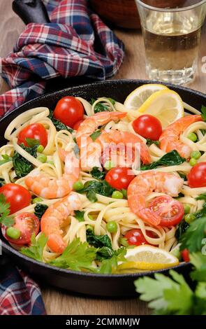 Pasta with fried prawns, peas, tomatoes and spinach in a frying pan, on a table with cider glasses. Stock Photo