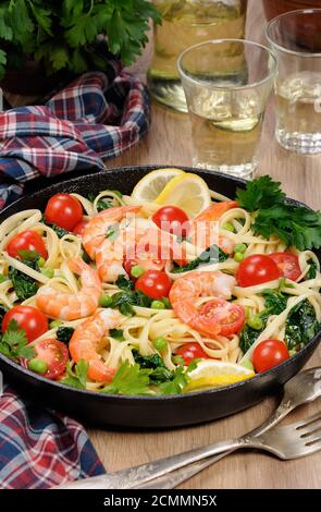 Pasta with fried prawns, peas, tomatoes and spinach in a frying pan, on a table with cider glasses. Stock Photo