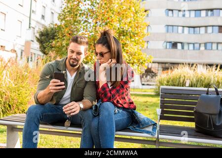 A couple being shocked over content seen on the screen of a phone while sitting during summer on a park bench in the city. Stock Photo