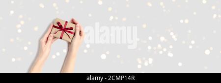 Female hands hold gift box with red bow on a gray background. Festive banner with golden shiny bokeh. Stock Photo