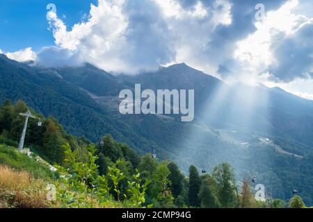 A ray of sun through the clouds illuminates the cable car in the mountains. Mountain Olympic Village, Krasnaya Polyana, Sochi, Russia. Stock Photo