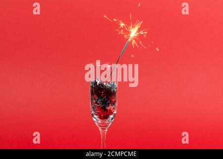 Glasses of champagne with confetti in shape of stars and sparkler on red background, Christmas decorations, copy space. Concepts of greeting card and Stock Photo