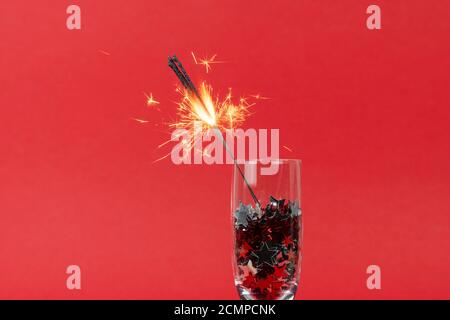 Glasses of champagne with confetti in shape of stars and sparkler on red background, Christmas decorations, copy space. Concepts of greeting card and Stock Photo