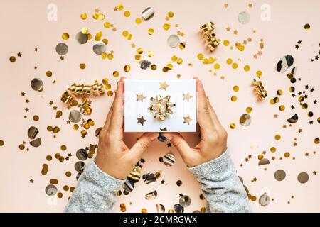 A girl holds a box with a gift in her hands on a festive pink background with gold confetti. Stock Photo