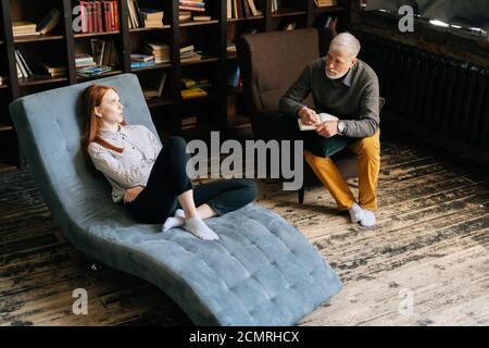 Attractive young woman on armchair talking about his problems with mature man psychotherapist Stock Photo