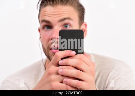 Macho with headphones listens to music. Man with surprised face holds mobile phone. Guy with beard holds phone or MP3 player and wears earphones on white background. Music and technology concept Stock Photo