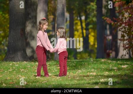 Two little sisters in identical clothes hold each other's hands during a walk in the park on a warm autumn day. Concept of a happy carefree childhood. Stock Photo