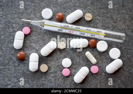 Colorful pills near to a medical mercury-in-glass thermometer indicating a critical temperature of 38.6 degrees Celsius on a dark gray table surface. Stock Photo