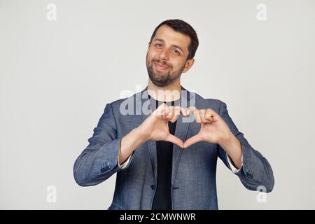 Young smiling businessman man with a beard in a jacket, smiling and making a heart shaped symbol with a finger, expressing love and positive romantic feeling. Portrait of a man on a gray background. Stock Photo
