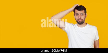 Young man with a beard in a white t-shirt worried about a problem, with a hand on his head, nervous and anxious about the crisis, stands on an isolated yellow background. Stock Photo
