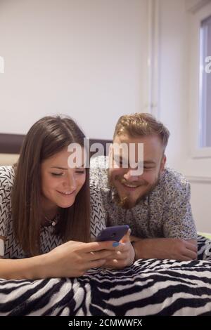 two people, couple or brother and sister, siblings laying together on bed, using phone together. Stock Photo