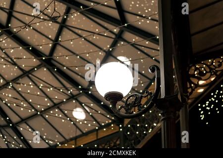 white round lantern on a wrought iron pillar of the restaurant terrace gazebo with garlands on the ceiling, architectural lighting of the room in a re Stock Photo