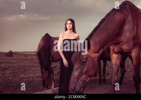Young woman in black dress at grassland sunset with horses herd Stock Photo