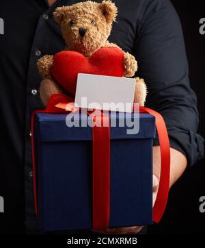 man in a black shirt holds a blue square box tied with a red ribbon and sits on top of a brown teddy Stock Photo