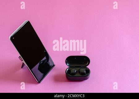 Wireless earphones, mobile phone on the phone stand, pink background. Modern technologies concept. Stock Photo