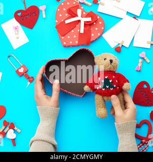 female hands holding a small brown teddy bear, on a blue background an open empty heart-shaped box Stock Photo