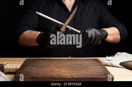 chef in a black shirt and black latex gloves sharpen a kitchen knife on an iron sharpener Stock Photo