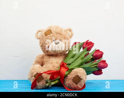 cute brown teddy bear with patches sits on a blue wooden background and holds in his paw a bouquet o Stock Photo