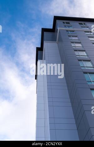 The corner top of a tall modern building with multiple colored grey metal panels. There's a blue sky with white clouds in the background. Stock Photo