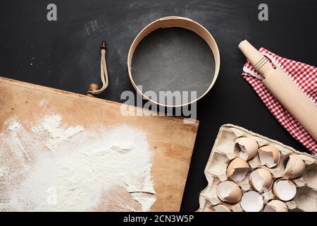wooden round sieve, white wheat flour, tray of chicken eggs in a tray and rolling pin on a black tab Stock Photo