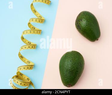 two green avocado fruits and a twisted measuring yellow tape on a beige-blue background Stock Photo