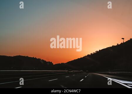 Roadtrip. Beautiful Sunrise with pink and blue clean sky on highway with smooth road and cars driving to the mountains silhouette through windshield. Stock Photo