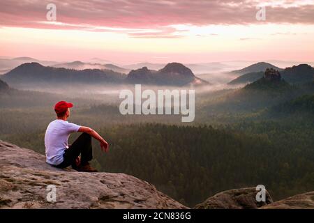 Adult sportsman in white shirt, dark trousars and red cap. Ginger short hair man sit on sharp cliff above valley in rocky mounta Stock Photo