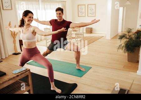 Couple exercising yoga indoors following instructions on laptop. Man and woman working out watching online fitness video tutorial. Stock Photo