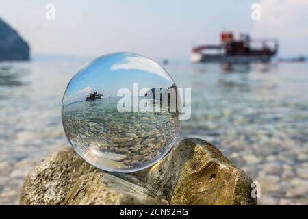 Crystal ball is on stones near the sea. Original upside down view and rounded perspective of the sky, sea and boat. Original and engaging picture. Stock Photo