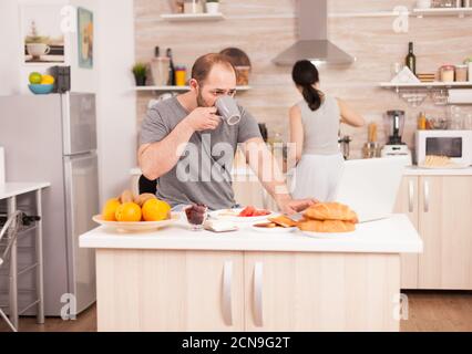 Freelancer drinking coffee and working from home while his wife prepares the breakfast. Freelancer work online via internet using modern digital technology Stock Photo