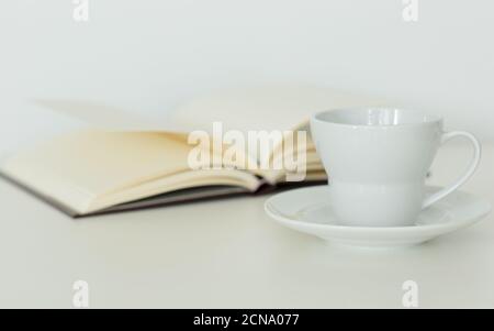 Open notebook with blank pages lying flat next to a coffee cup on a saucer. Isolated against white background. Calm study and relaxation. Stock Photo