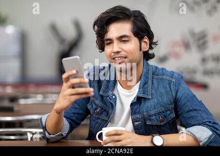 Portrait of a young handsome Indian man checking his phone, sitting in an office cafeteria, coffee shop, casual work environment. Stock Photo