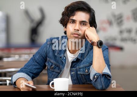 Portrait of a young handsome Indian man with serious look, lost in thought, sitting in an office cafeteria, coffee shop, casual work environment. Stock Photo