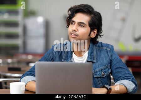 Portrait of a young handsome Indian man looking away from his laptop, sitting in an office cafeteria, coffee shop, casual work environment. Stock Photo