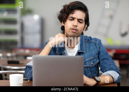 Portrait of a young handsome Indian man working on his laptop, sitting in an office cafeteria, coffee shop, casual work environment. Stock Photo