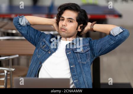 Portrait of a tired Indian man working on his laptop, sitting in an office cafeteria, coffee shop, casual work  head resting back, lost in thought. Stock Photo